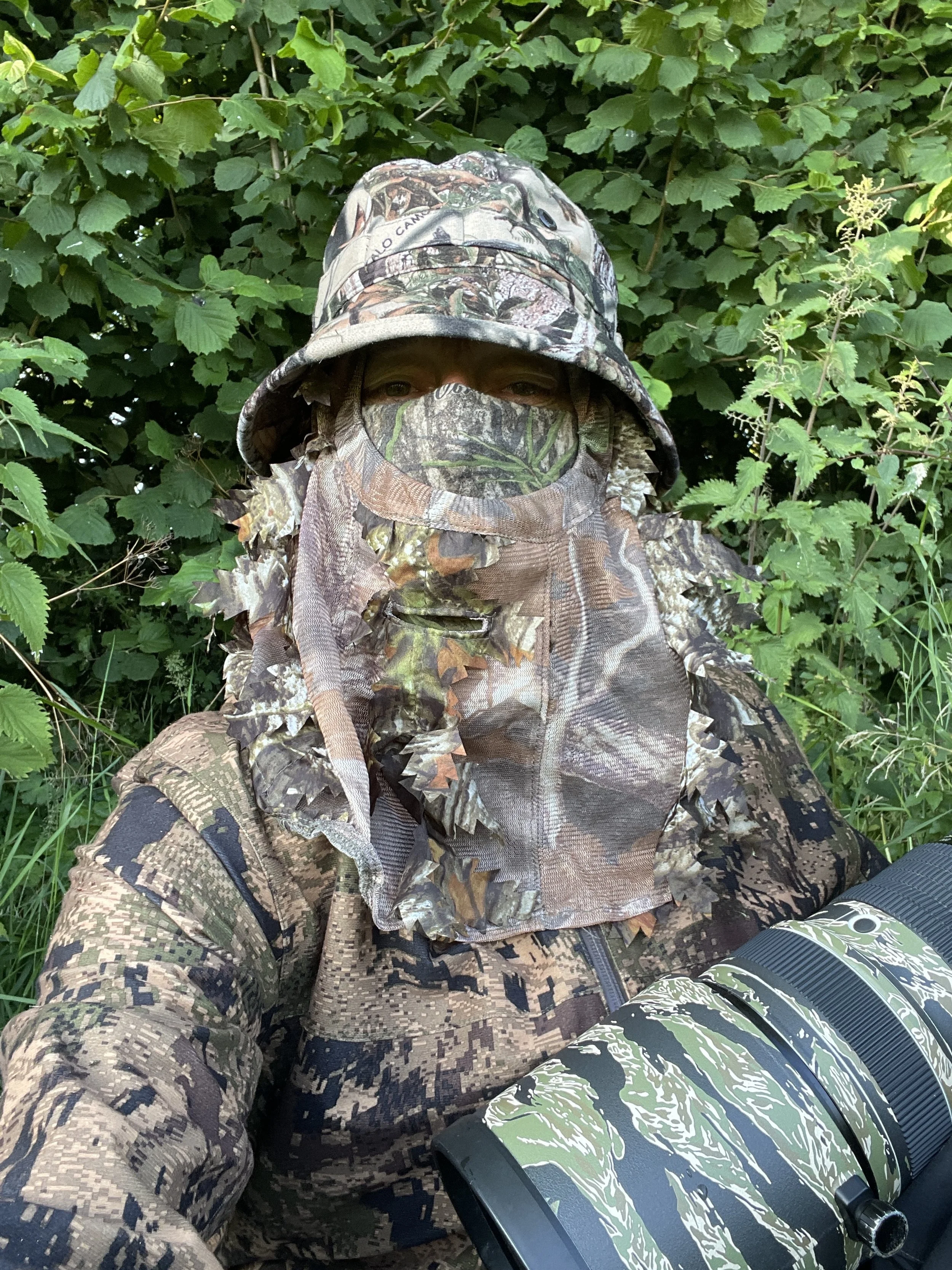 Person dressed in camouflage hunting gear with a hat and face covering, standing in a green, leafy outdoor setting with a camouflage-patterned telescope in front.