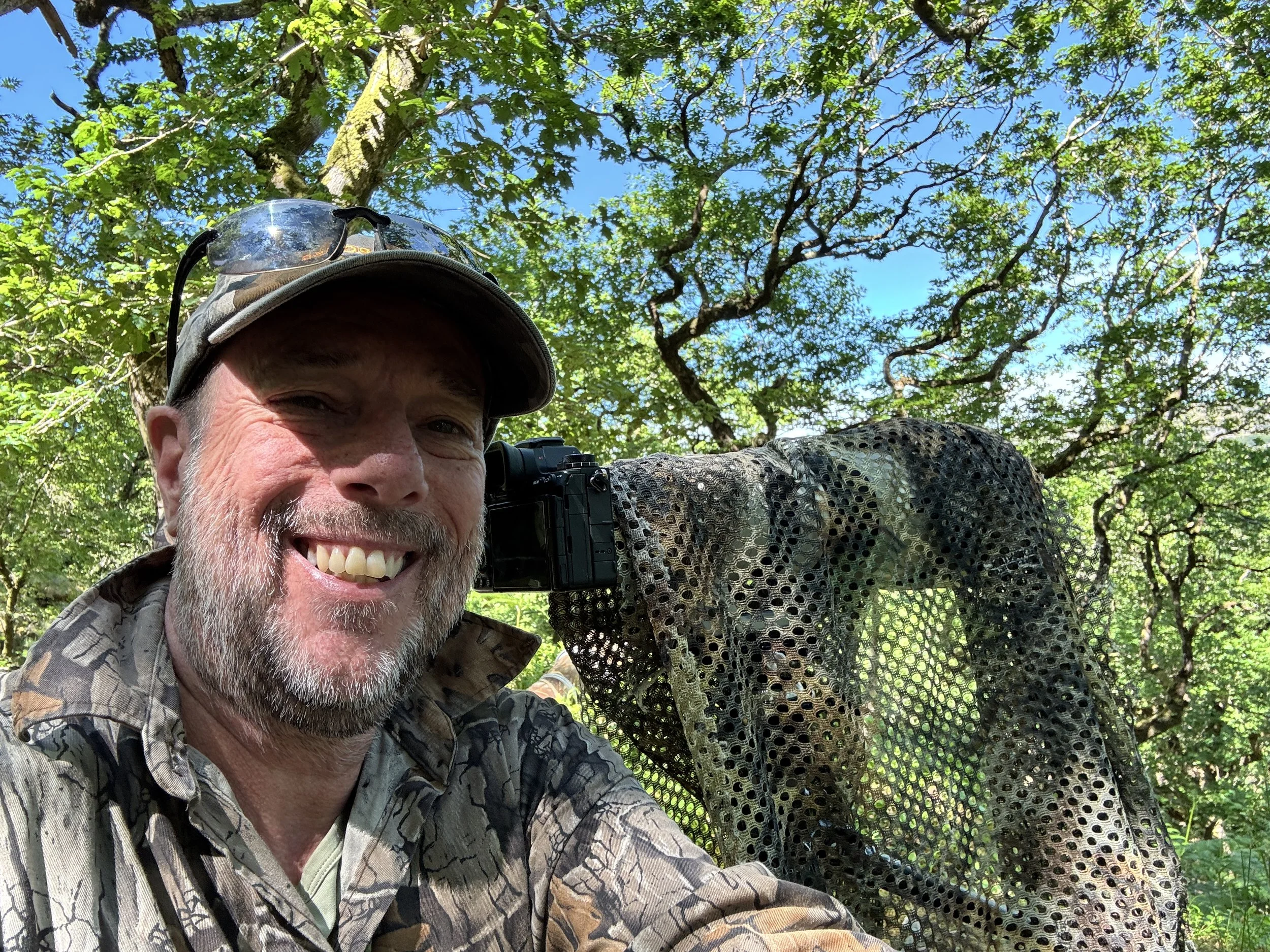 A smiling man wearing a camo jacket and cap with glasses on it, taking a selfie outdoors in a lush green forest, with a camera set up with a black mesh cover behind him.