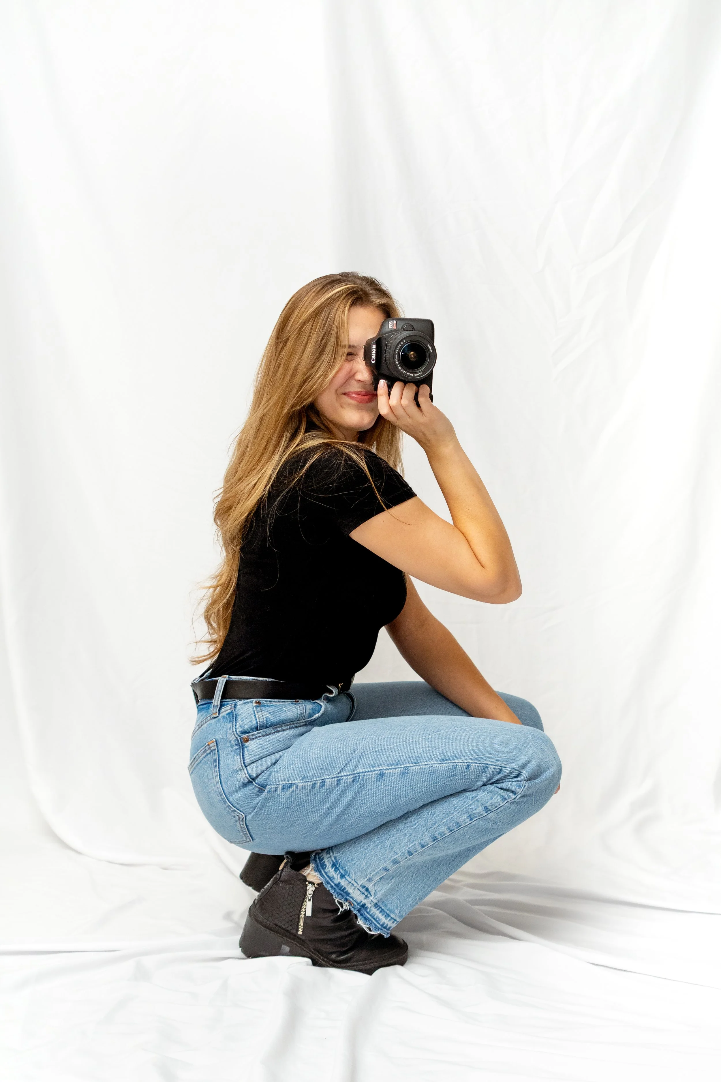 A woman with long blonde hair squatting and taking a selfie with a camera in front of a white backdrop.