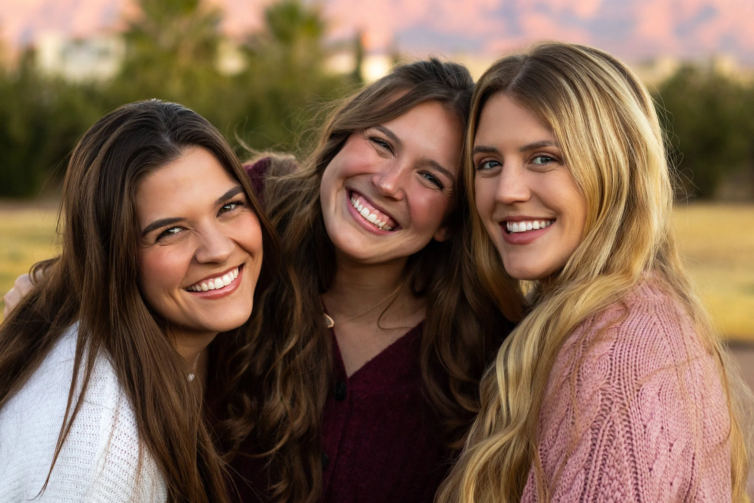 Three smiling women outdoors close together with blurred trees and mountains in the background.