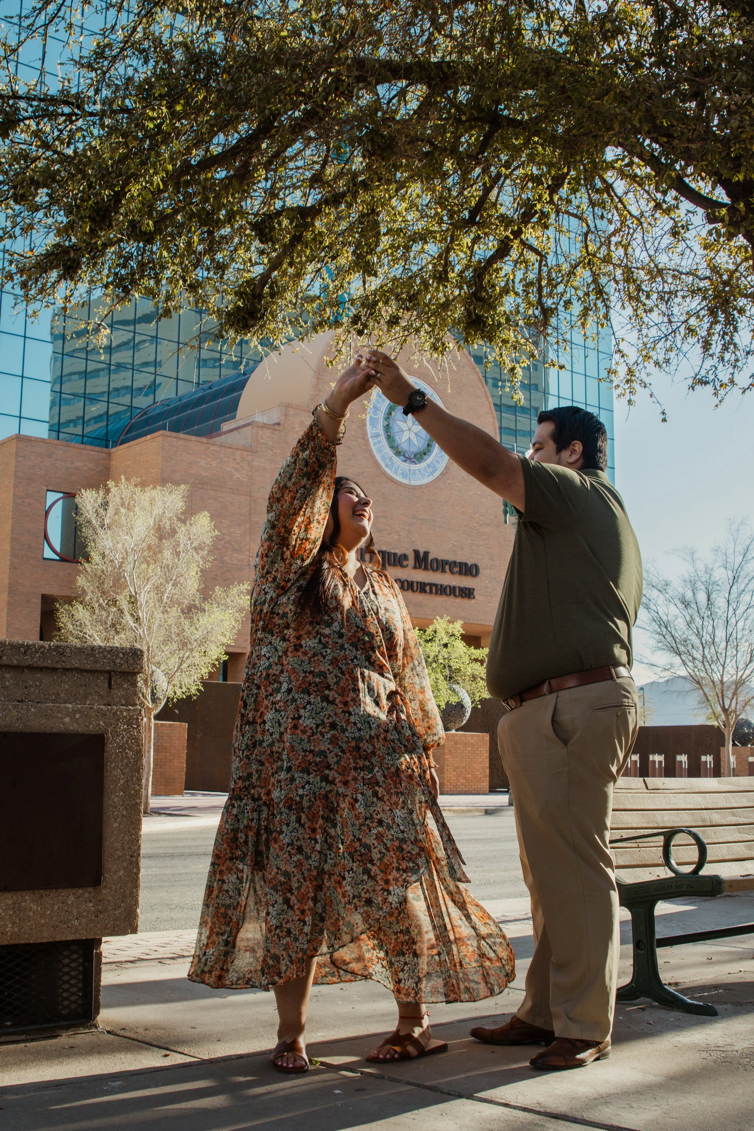 A couple dancing outside in front of a modern courthouse building with their hands raised together, smiling and enjoying the moment on a sunny day.