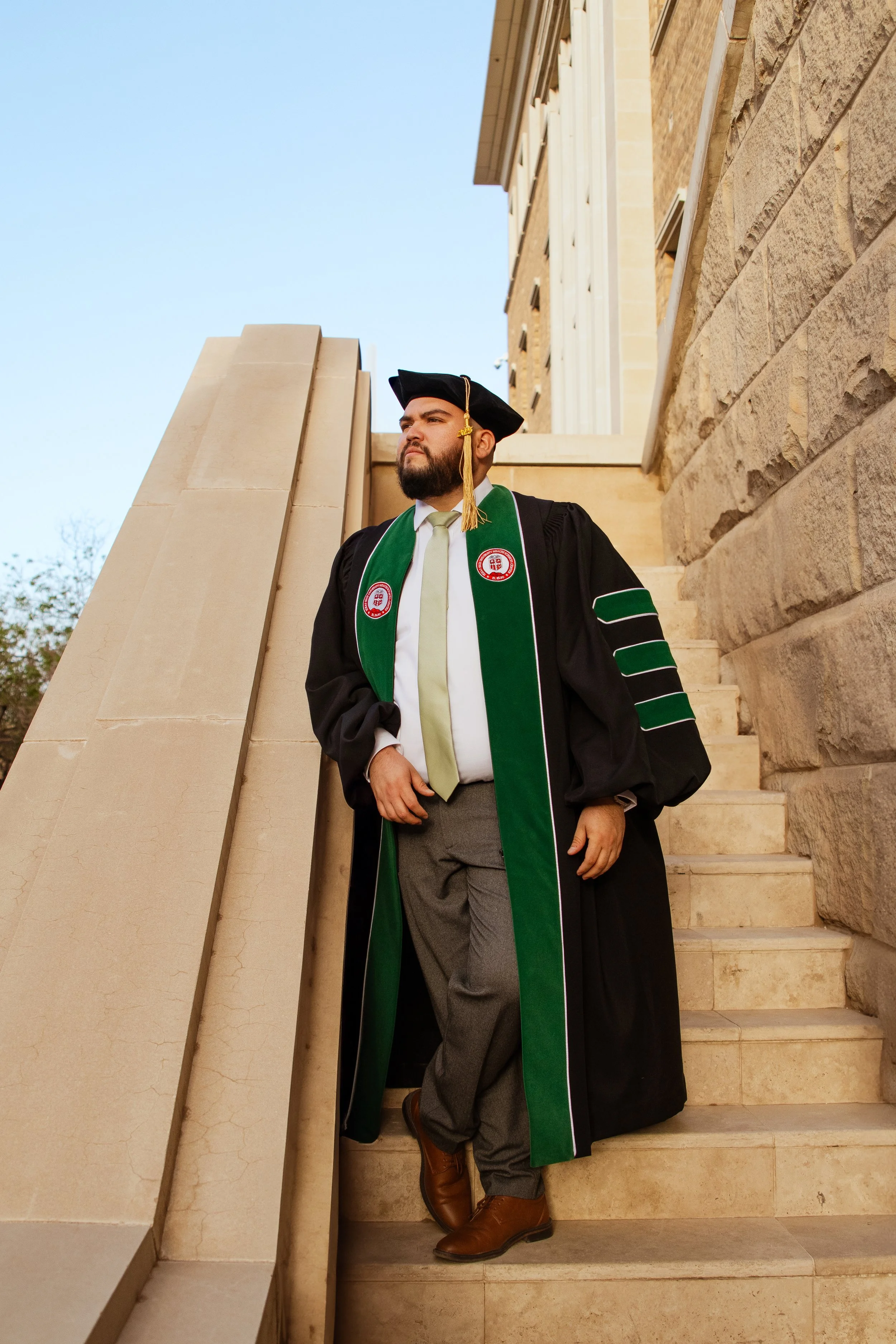 A man in graduation attire, including a black gown, green stole with red and white emblems, and a black cap with a gold tassel, standing on stone steps outside a building, looking to the side.