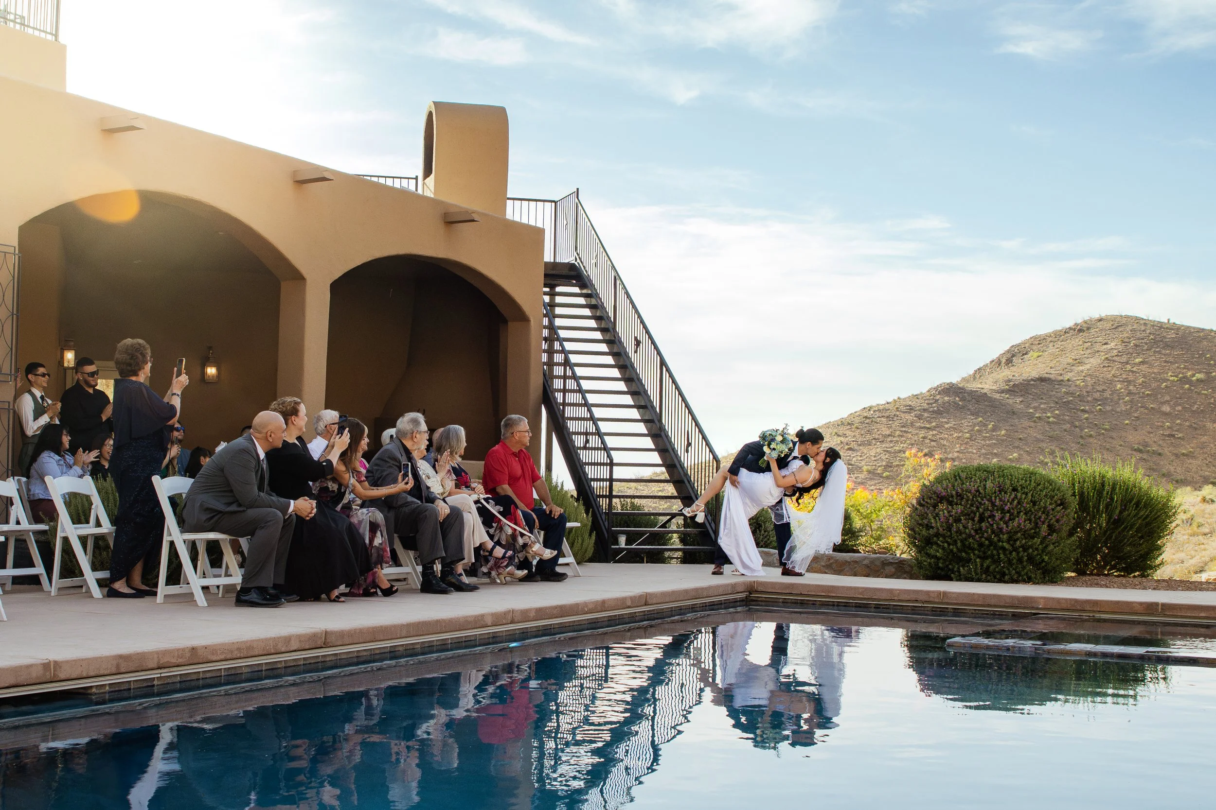 A wedding ceremony outdoors by a pool in a desert landscape, with guests seated and a couple sharing a kiss.