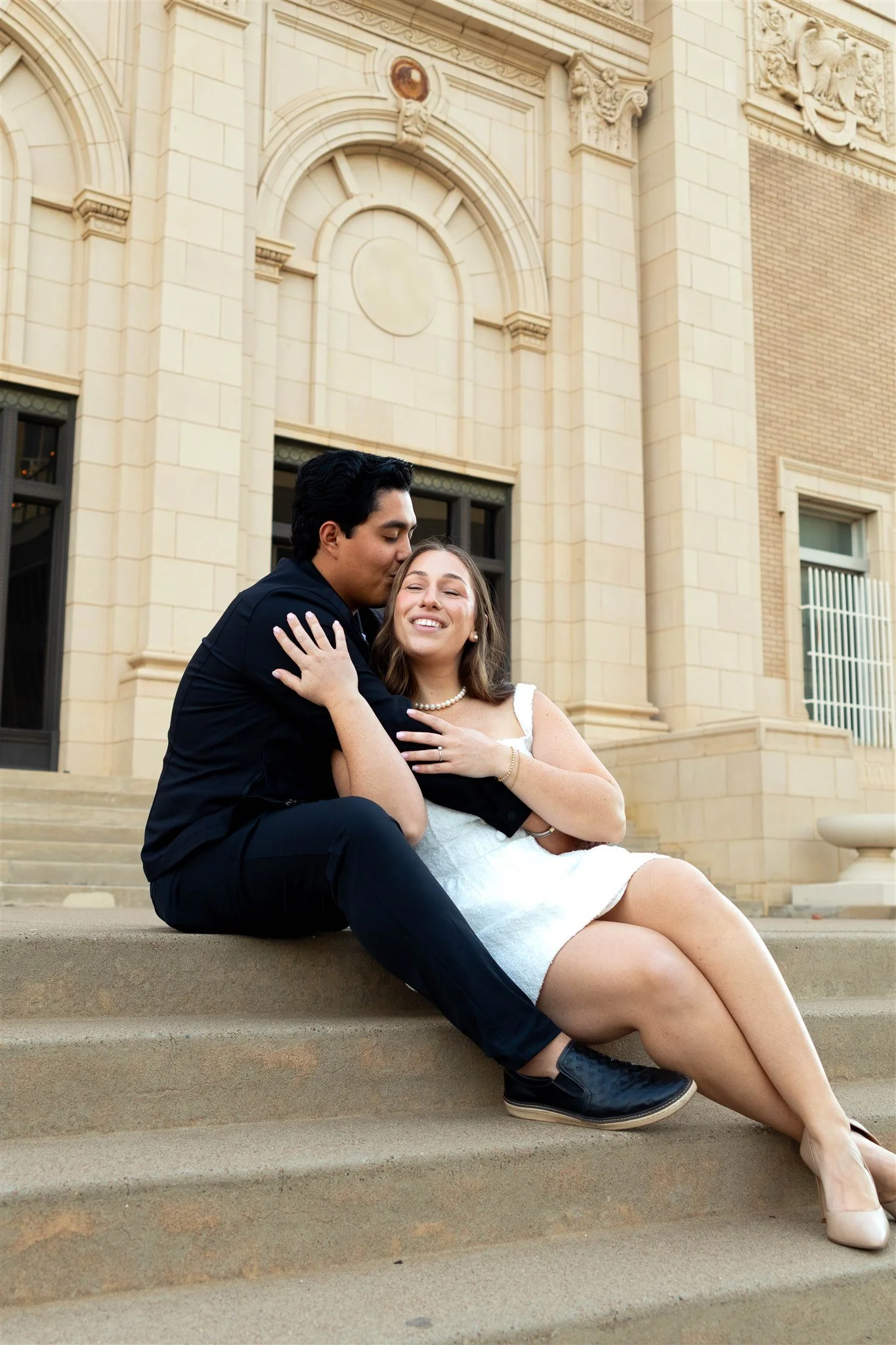 A couple sitting on steps in front of a historic building, sharing a tender moment with the man kissing the woman's temple and the woman smiling with closed eyes, dressed in formal attire.