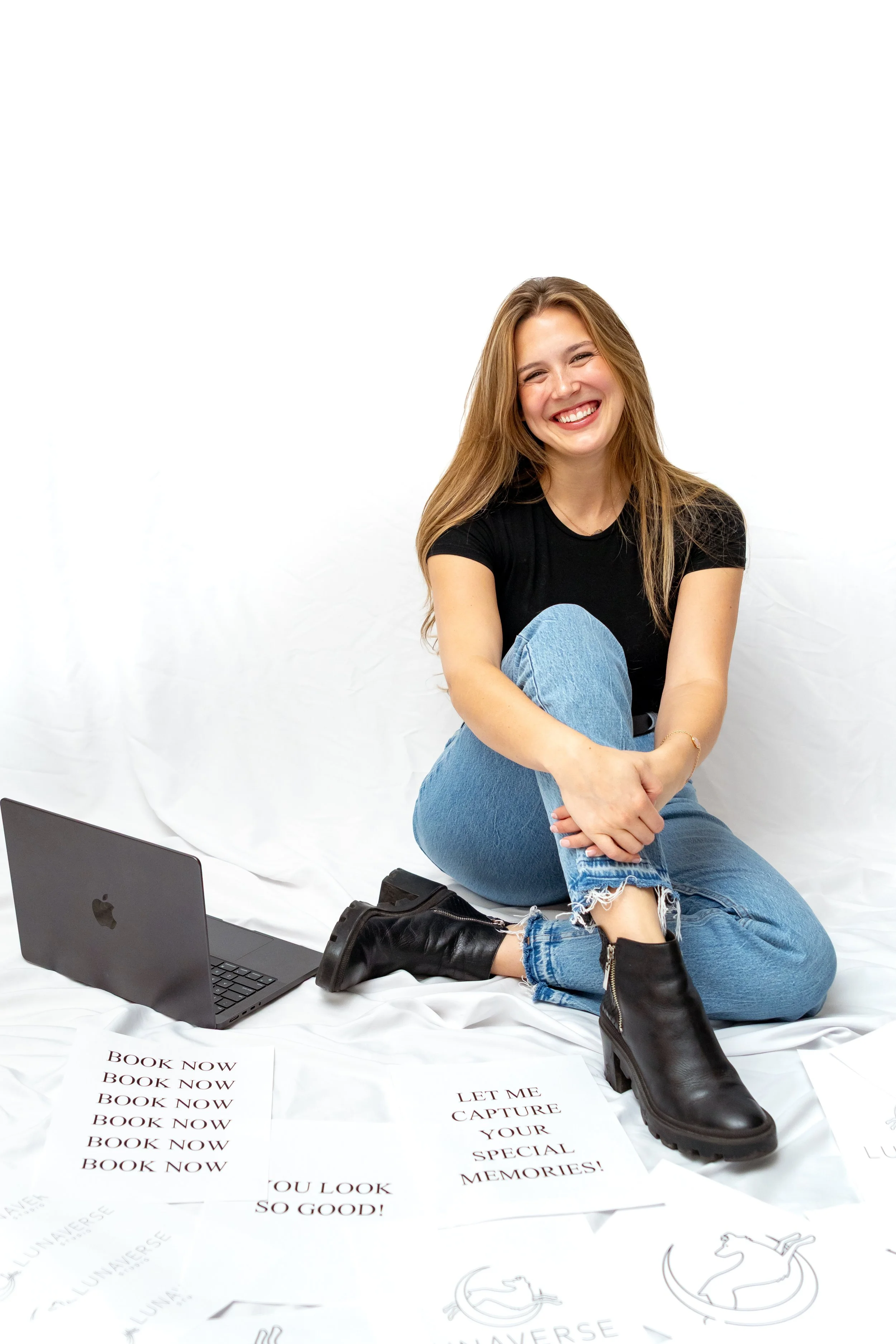 A smiling young woman sitting on a white backdrop, wearing black boots, blue jeans, and a black t-shirt, surrounded by printed papers and a laptop.