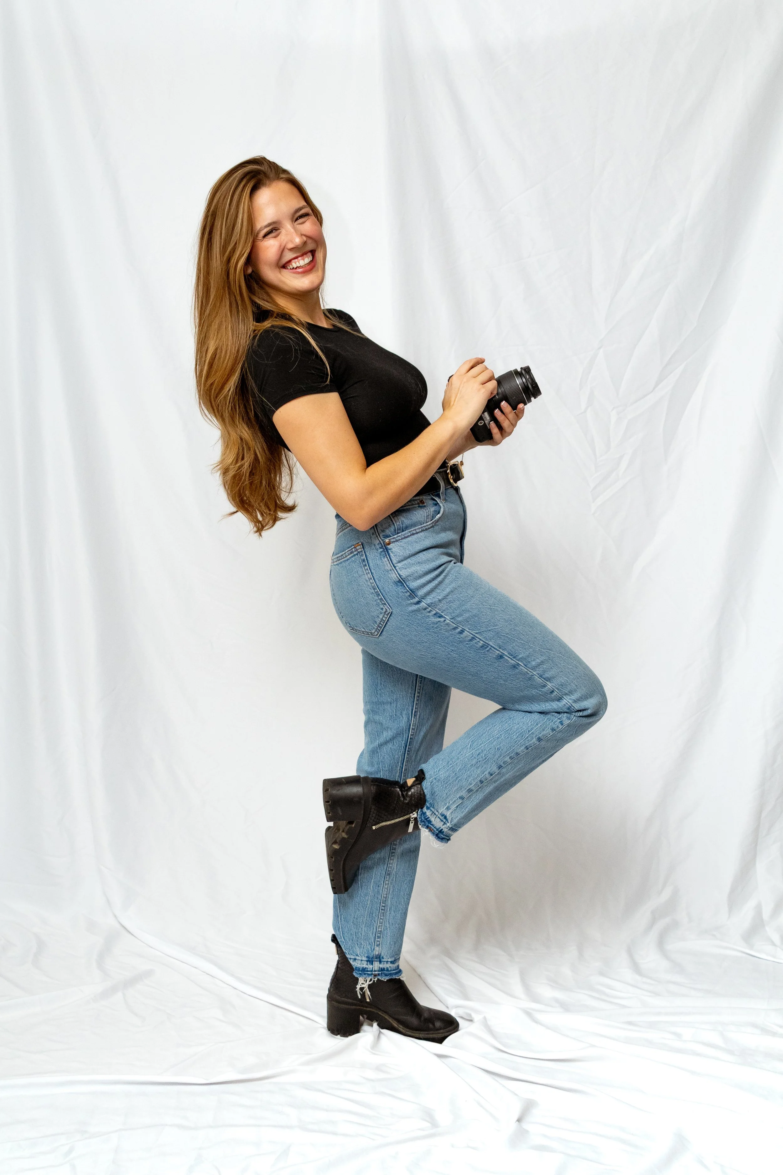 A woman with long brown hair wearing a black t-shirt, blue jeans, and black boots, holding a camera, standing on one leg against a white backdrop, smiling.