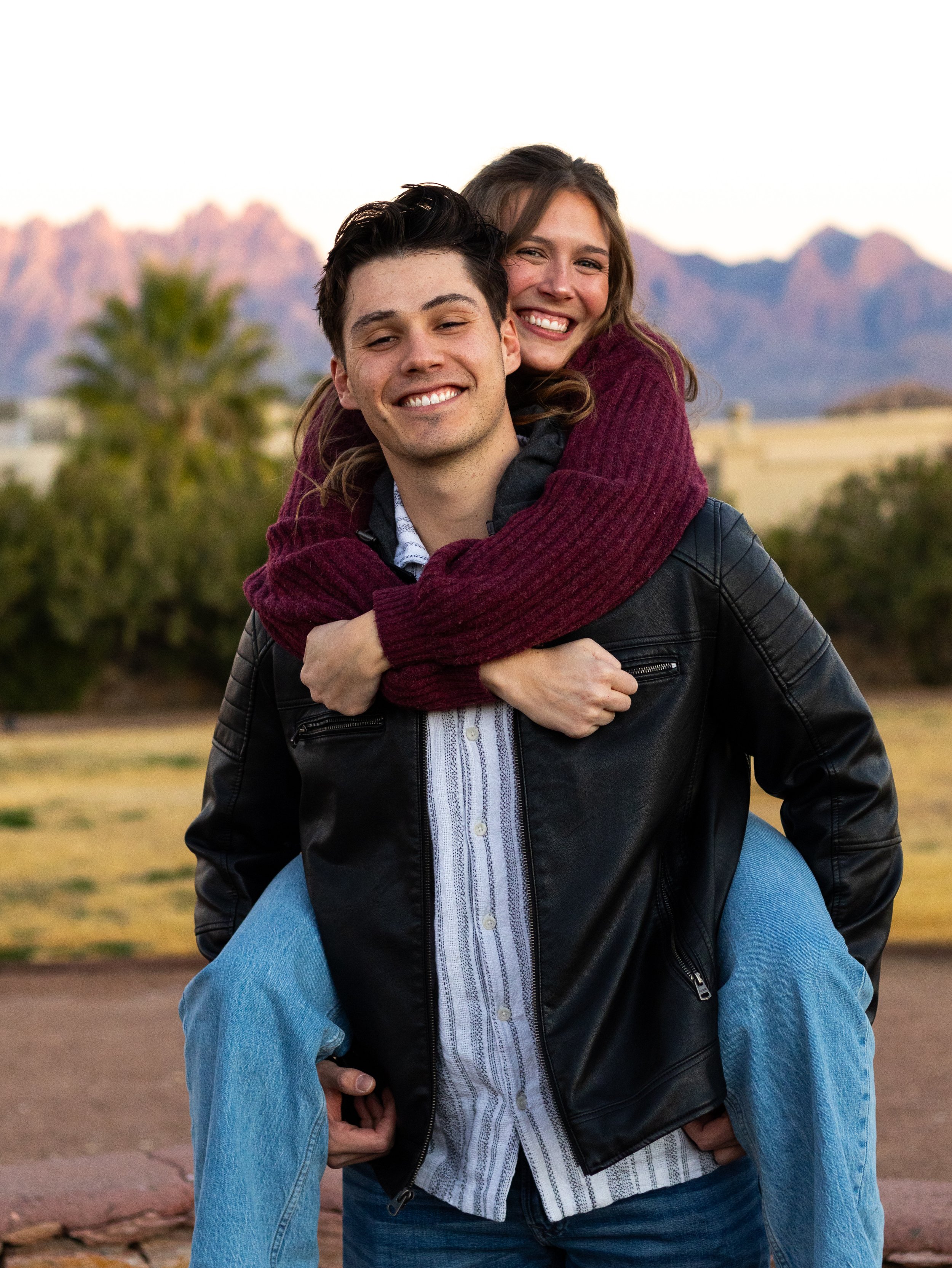 A smiling young man giving a piggyback ride to a laughing young woman. They are outdoors with mountains in the background, early evening lighting.