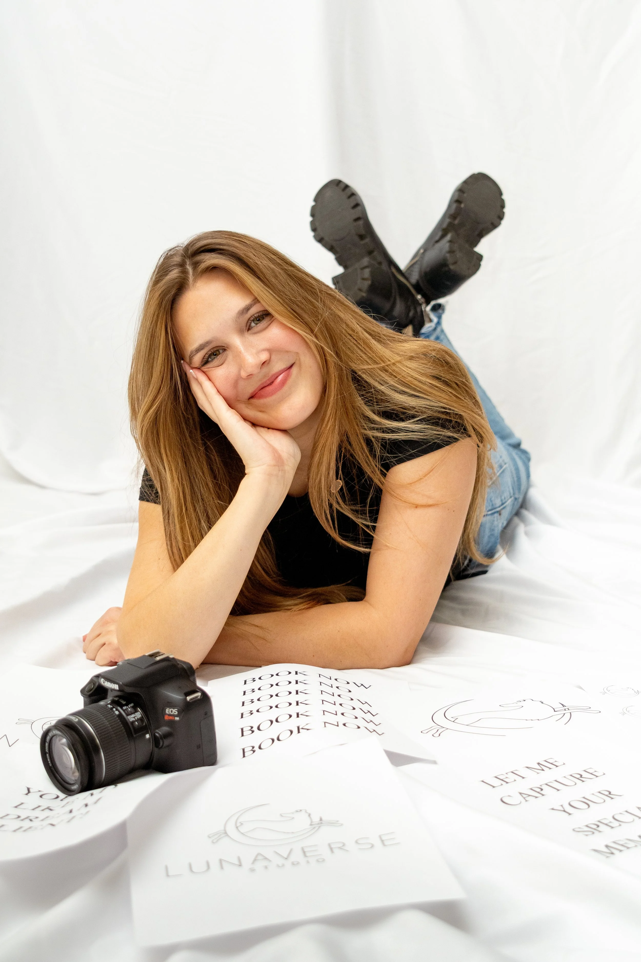 A young woman with long reddish-brown hair lying on a white surface, smiling and resting her head on her hand. She wears a black t-shirt, jeans, and black boots, with a Canon camera in front of her. Sheets of paper with logo and text are spread out in front of her, some with the word 'BOOK' and others with the phrase 'LET ME CAPTURE YOUR'. The background is a plain white backdrop.