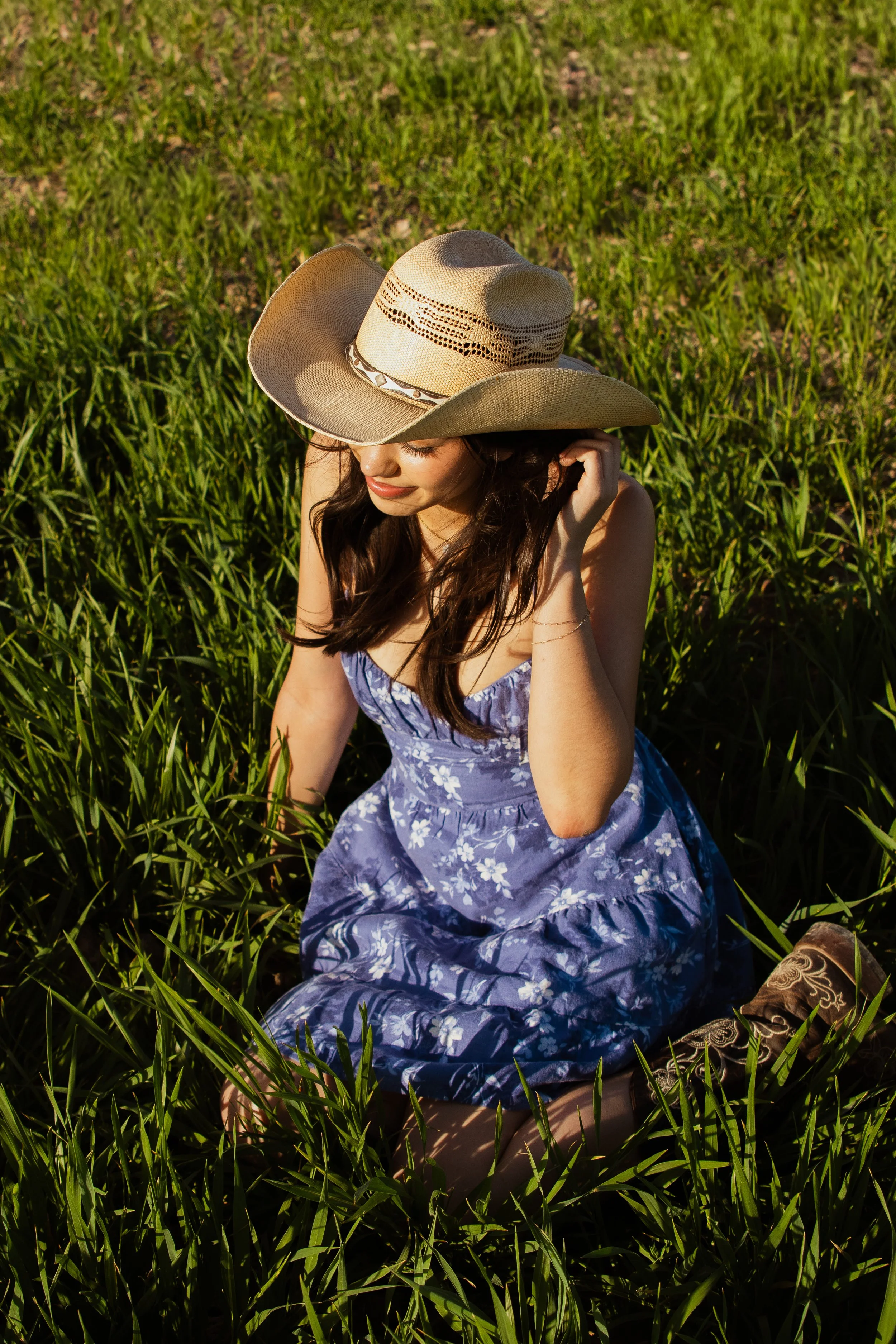 A woman sitting in tall green grass, wearing a blue floral dress and a large straw hat, with her eyes closed and a peaceful expression.