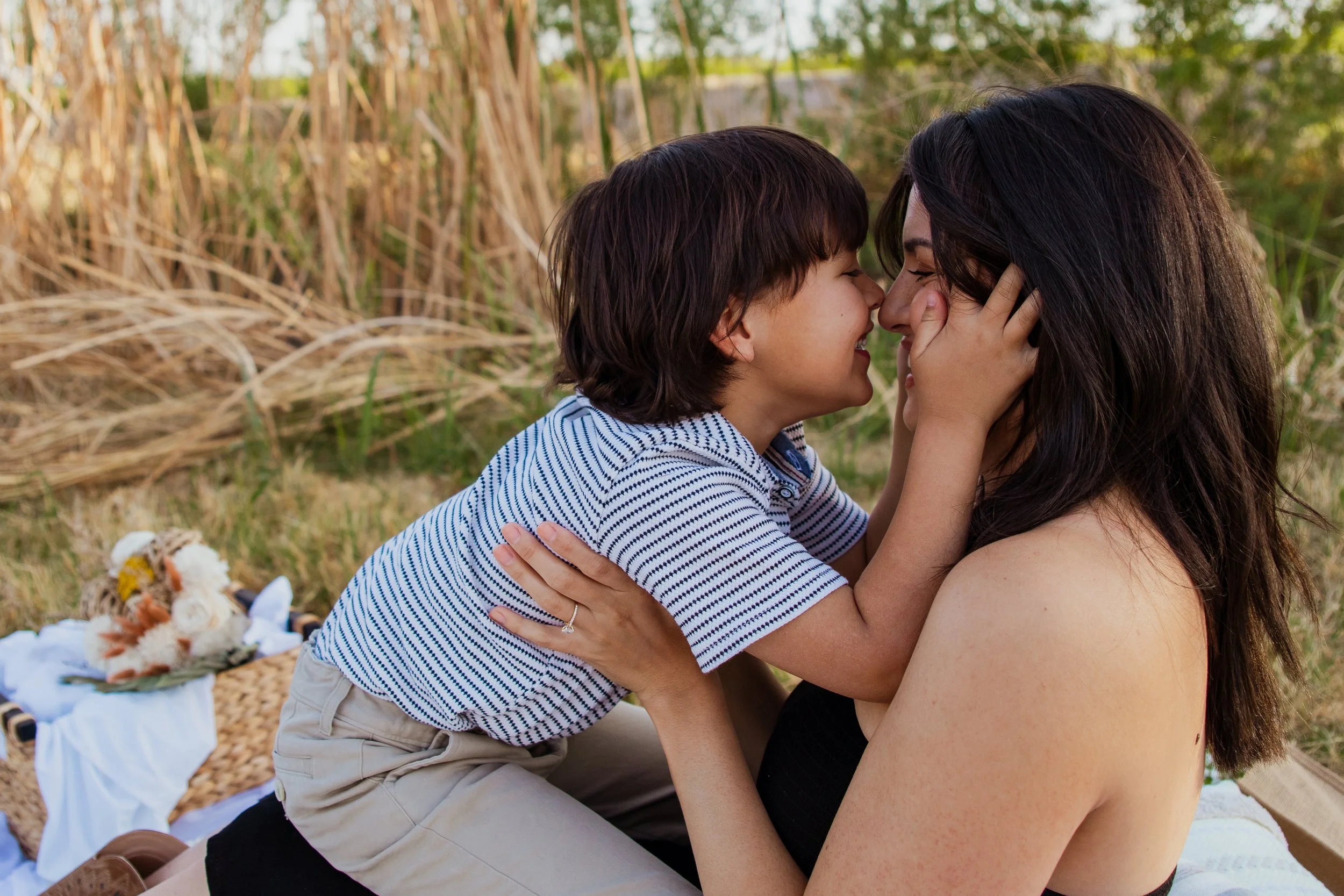 A woman and young boy share a close, affectionate moment outdoors in a grassy area with tall, dry grass in the background, smiling and touching noses.