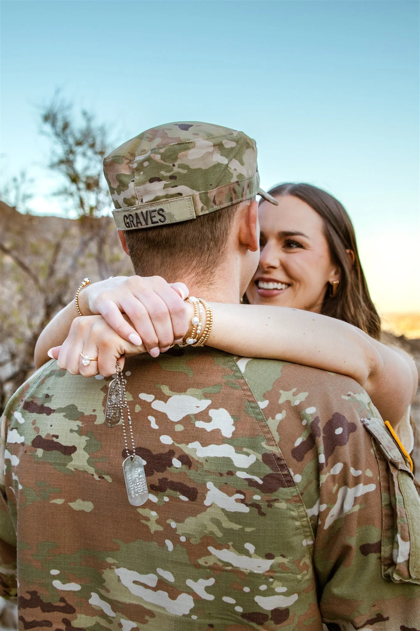 A soldier in camouflage uniform and cap with 'GRAVES' on it is embracing a smiling woman, with her arms around his neck, outdoors during sunset.