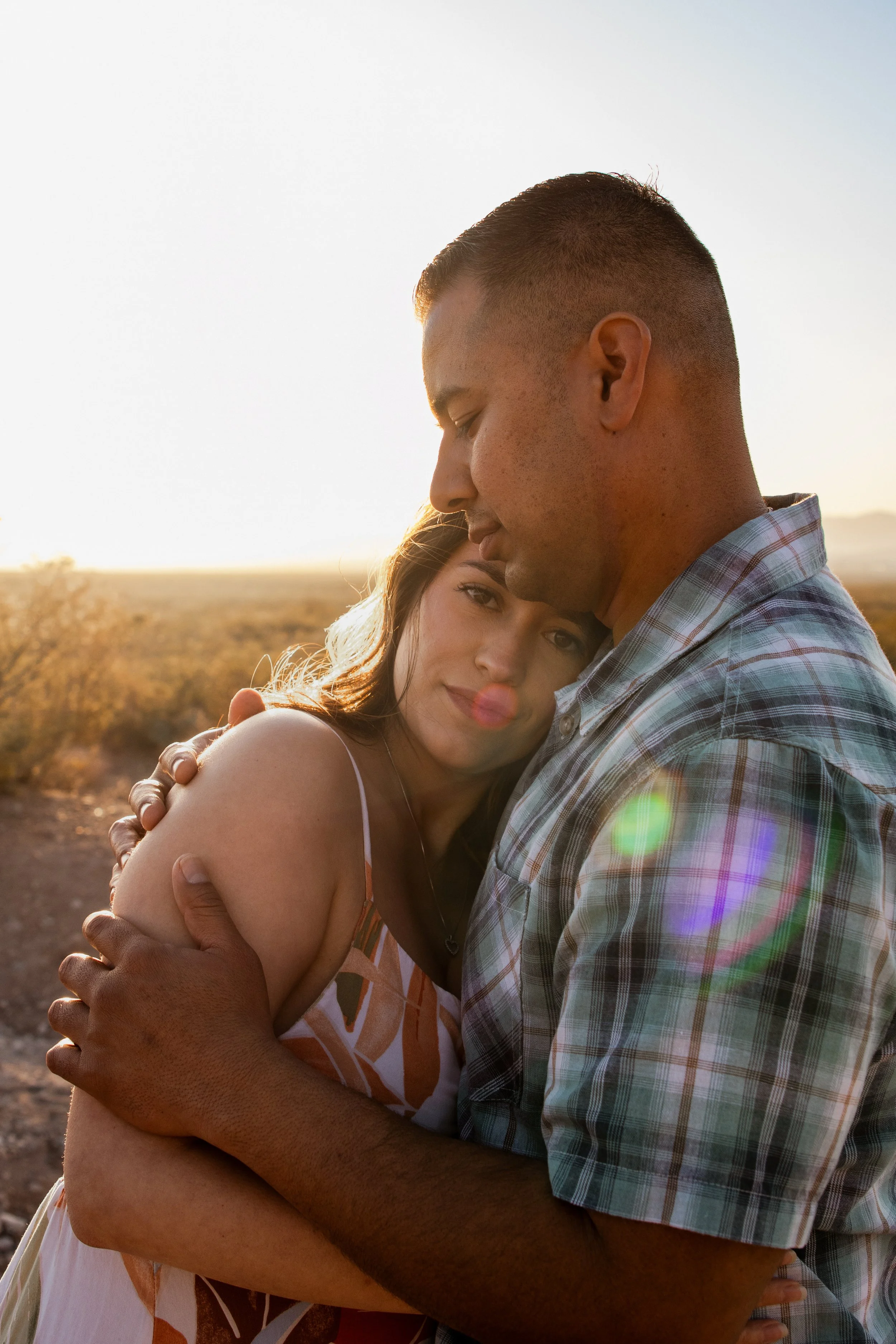 A man and woman hugging outdoors during sunset, their faces close together, with soft sunlight and a landscape in the background.
