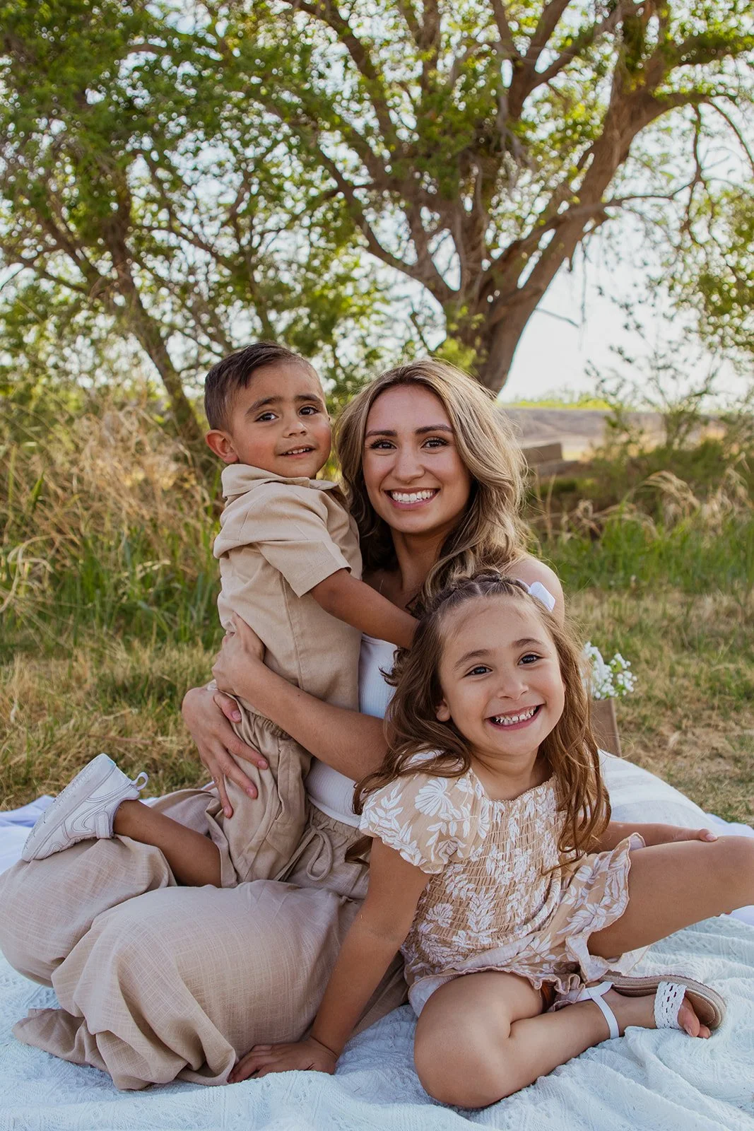 A woman with two children sitting on a blanket outdoors under a tree, smiling at the camera. The woman is in the middle, the girl with curly hair is sitting in front, and the boy with short hair is sitting behind her.