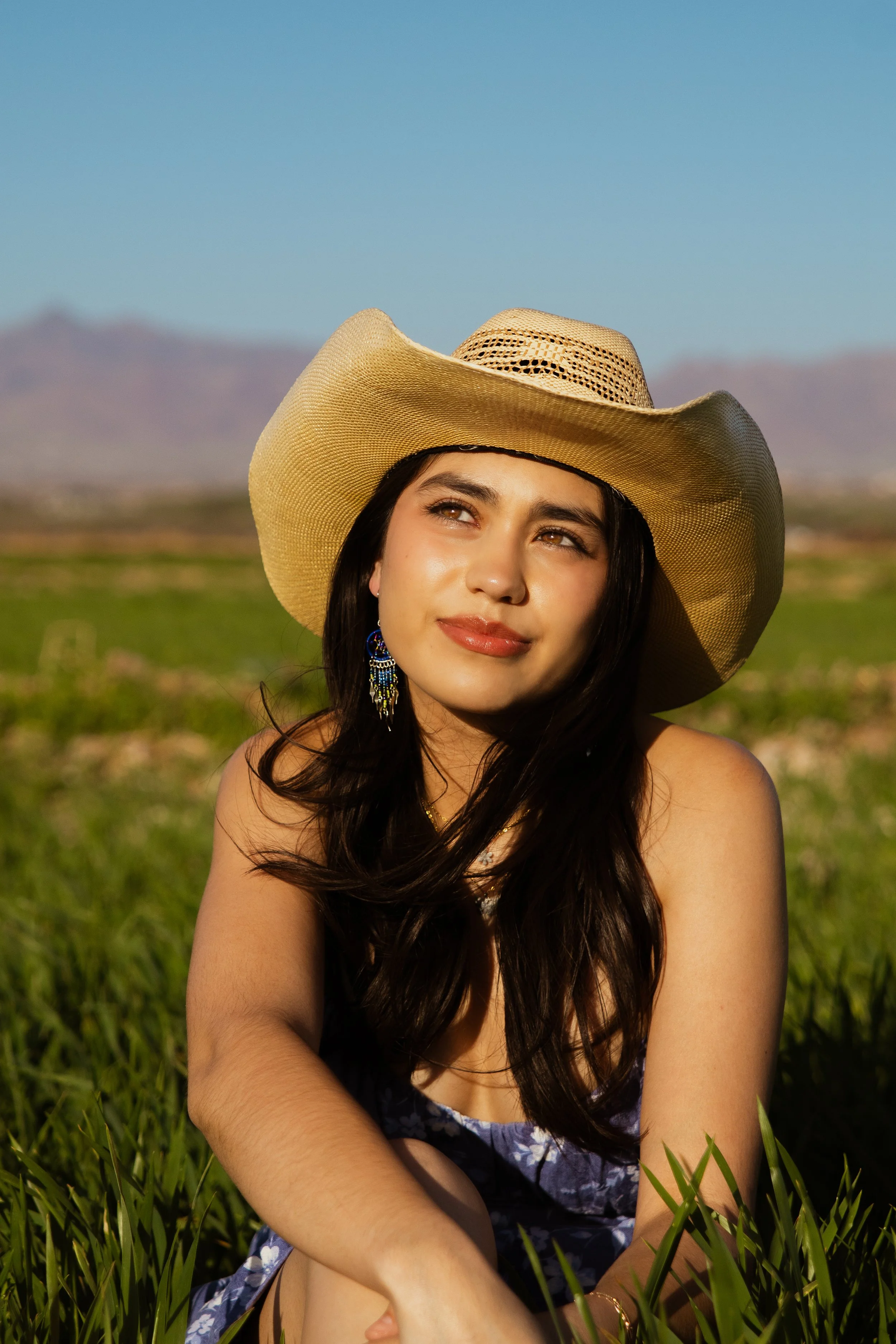 A woman sitting in a field of grass wearing a wide-brimmed straw hat and a floral dress, with mountains in the background and clear blue sky.