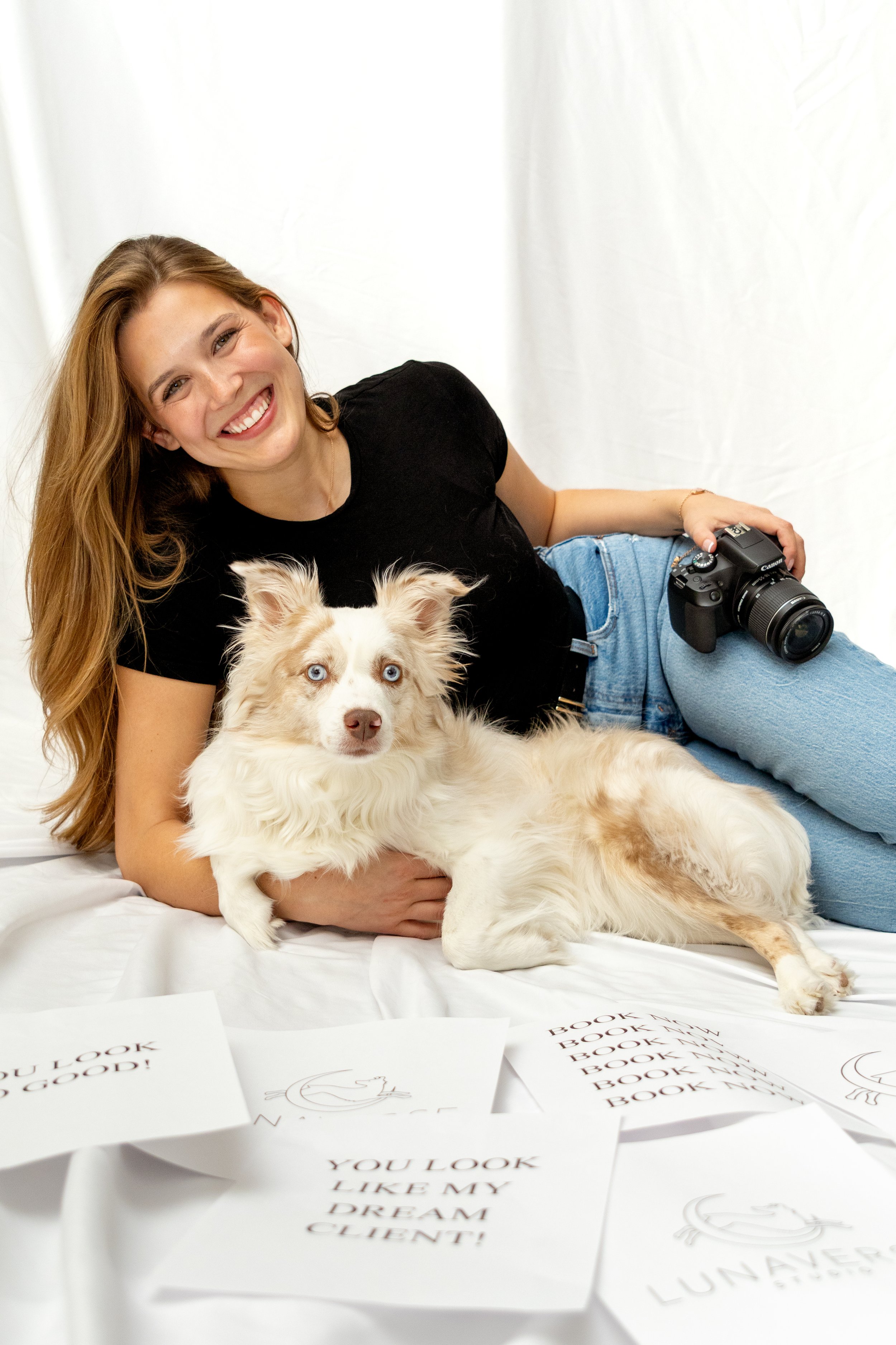 A woman with long red hair smiling, holding a white and tan Australian Shepherd dog with blue eyes, lying on white fabric with printed papers around them.