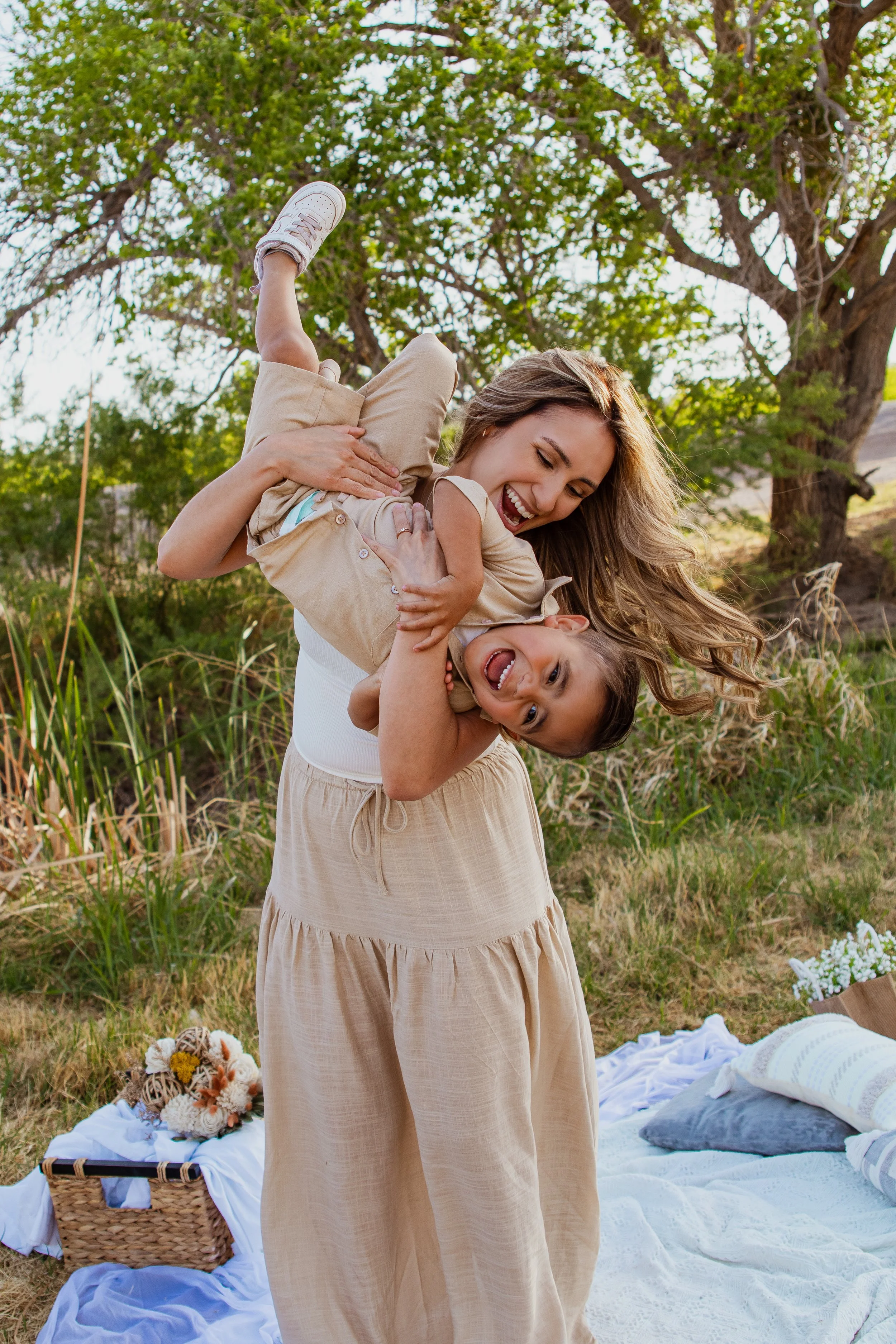 A woman and a young girl are outdoors, laughing and playing. The woman is holding the girl upside down, and they are both smiling and having fun on a grassy area with trees, a picnic blanket, and picnic items nearby.