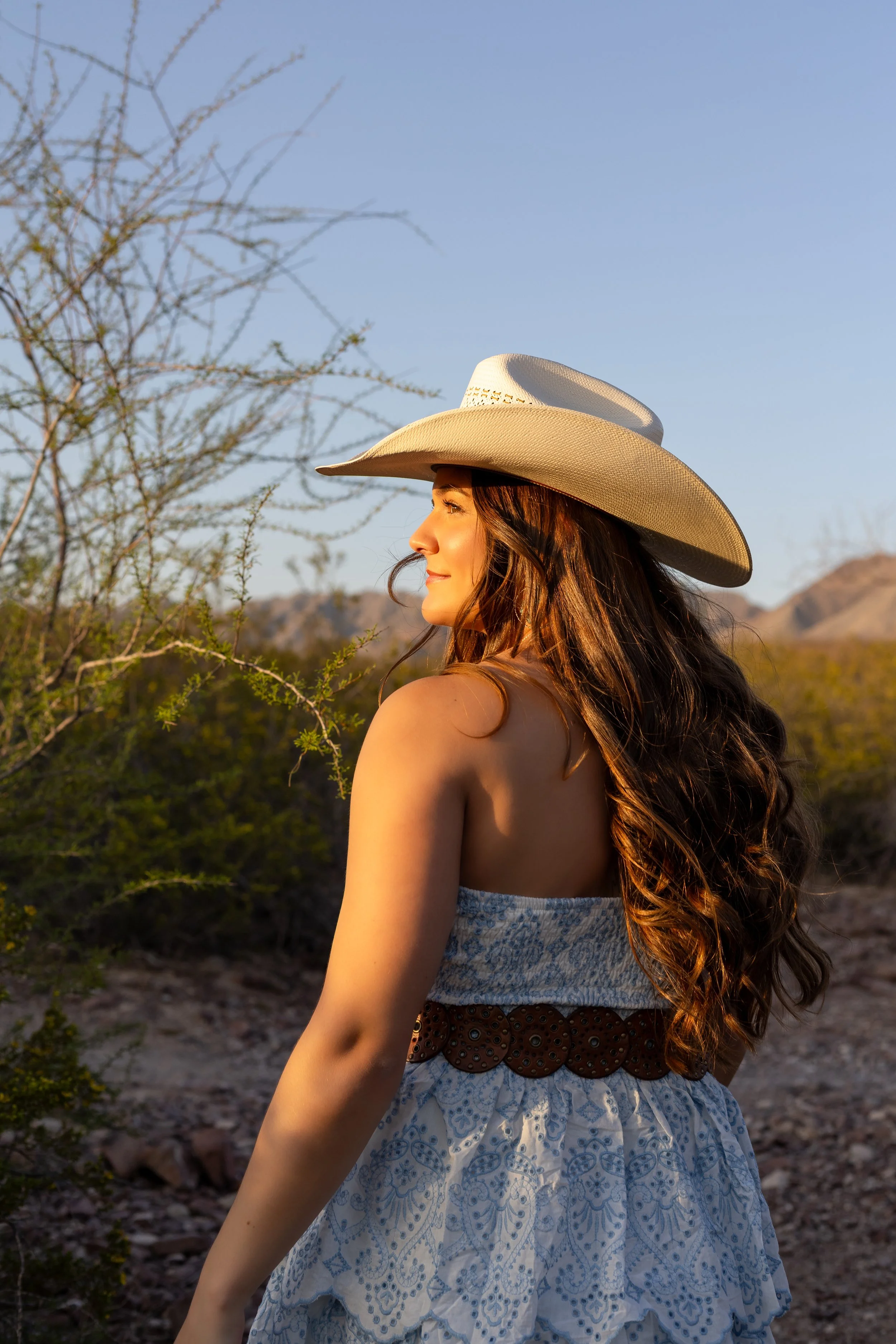 A woman with long wavy hair wearing a wide-brimmed cowboy hat and a strapless summer dress, standing outdoors in a desert landscape during daytime.