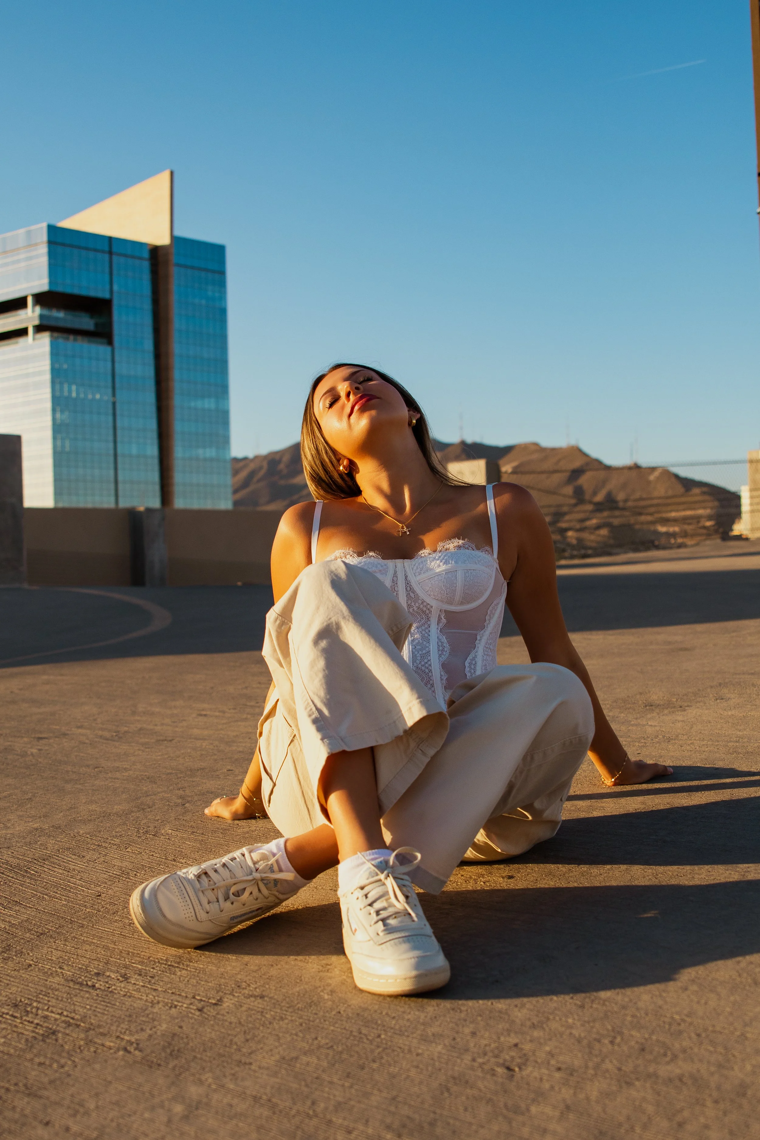 Young woman sitting on a rooftop parking lot in the late afternoon sunlight, with modern buildings and mountains in the background.