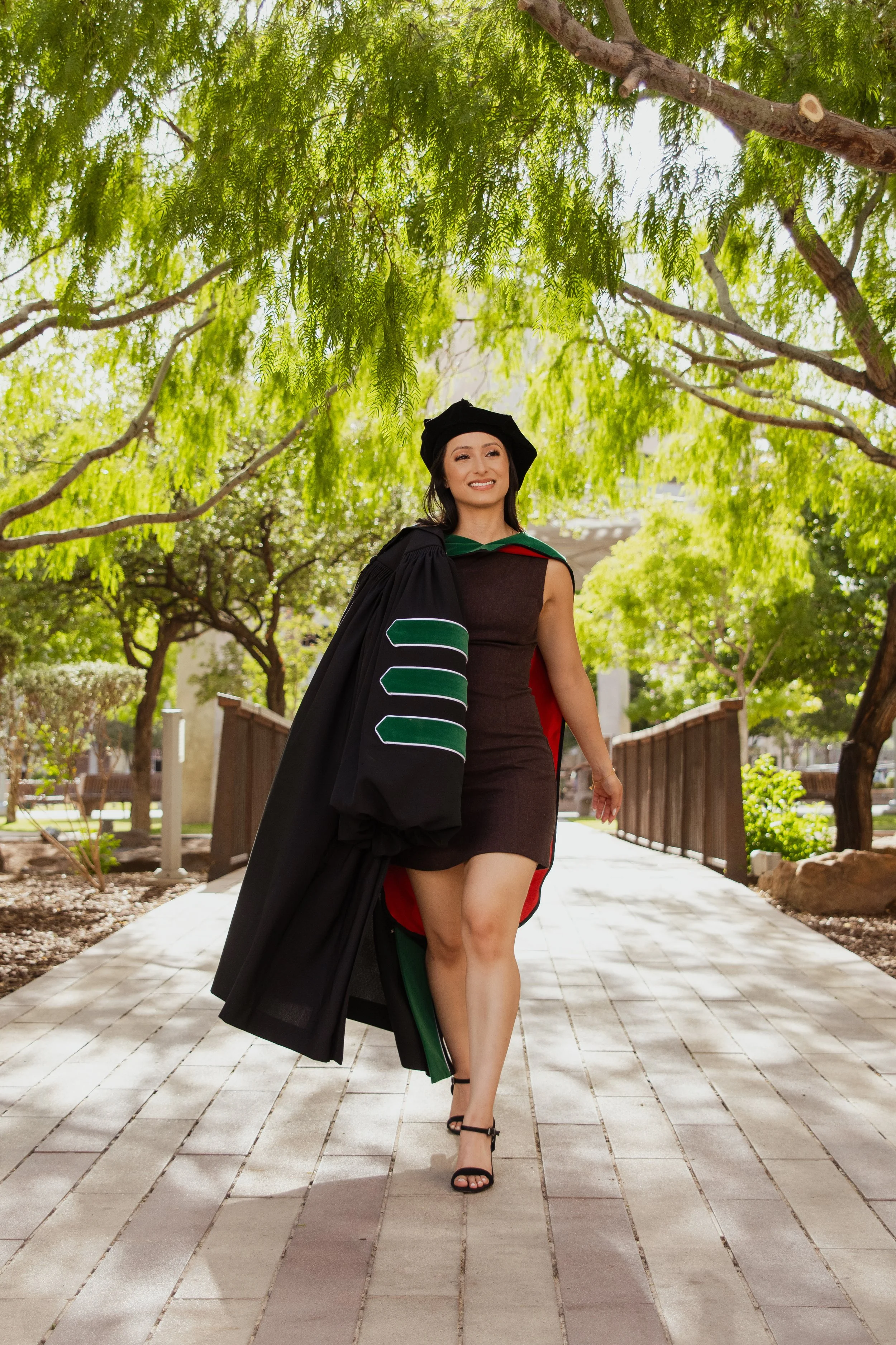 A woman in a graduation gown and cap walking outdoors on a paved walkway surrounded by green trees.