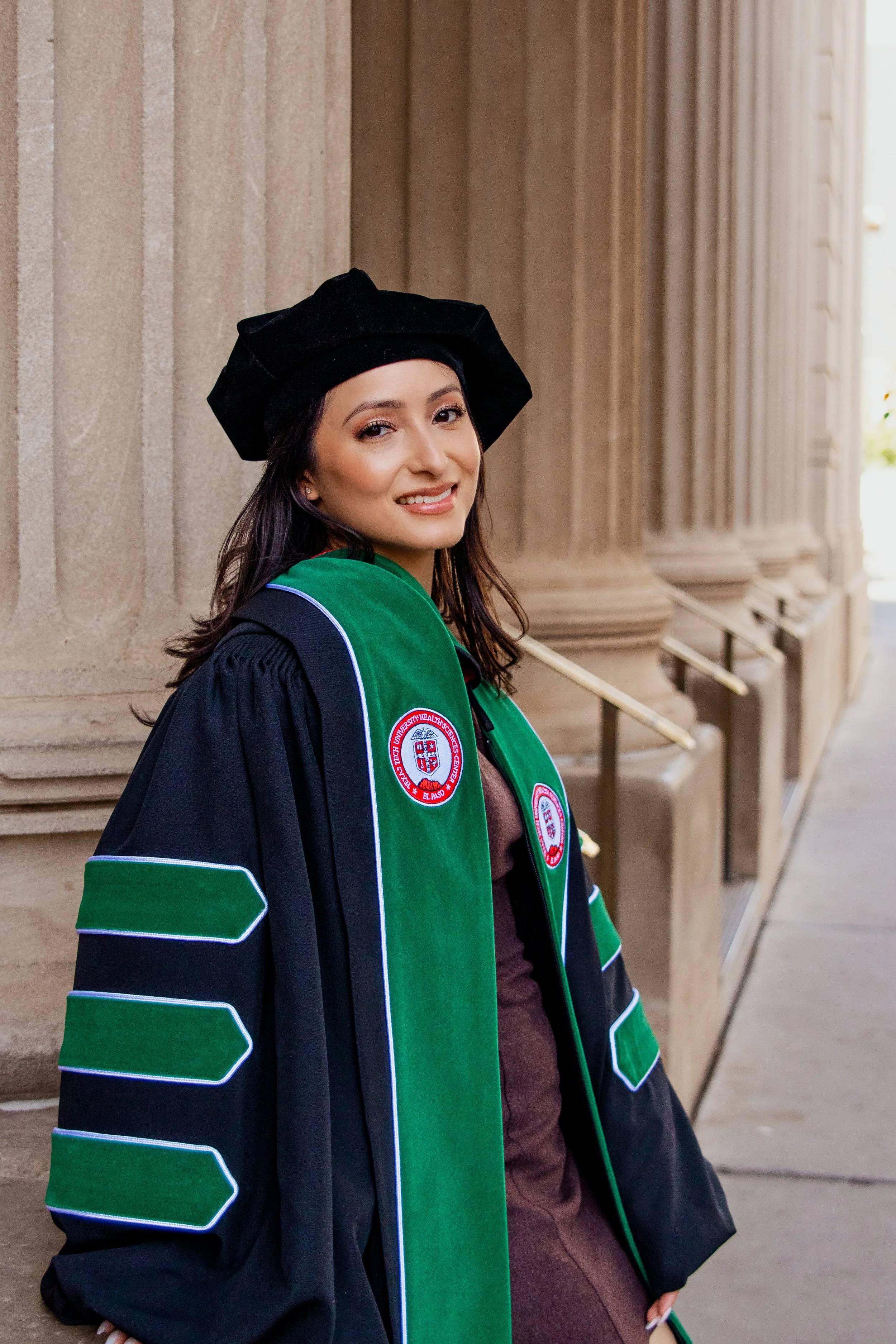 A young woman in graduation attire, wearing a black cap and gown with green and black academic regalia, standing outdoors in front of classical stone columns, smiling.