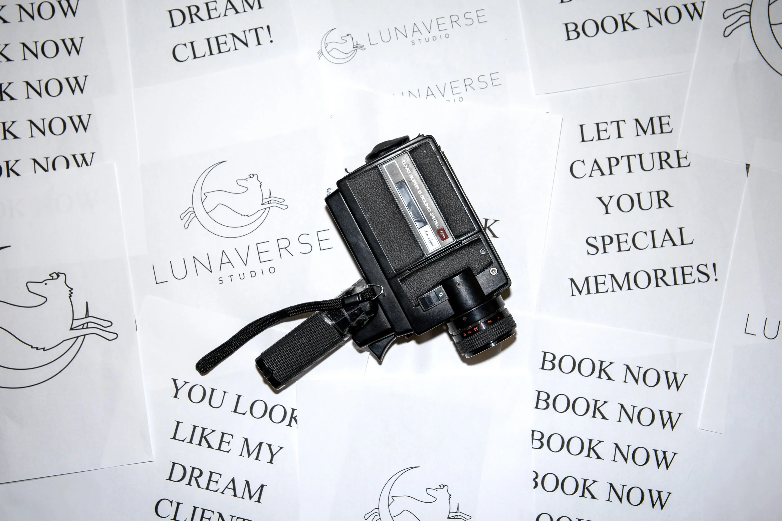 A vintage video camera resting on a collection of printed advertising sheets for LunaVerse Studio, with phrases like "Book Now," "Let me capture your special memories," and "You look like my dream client."