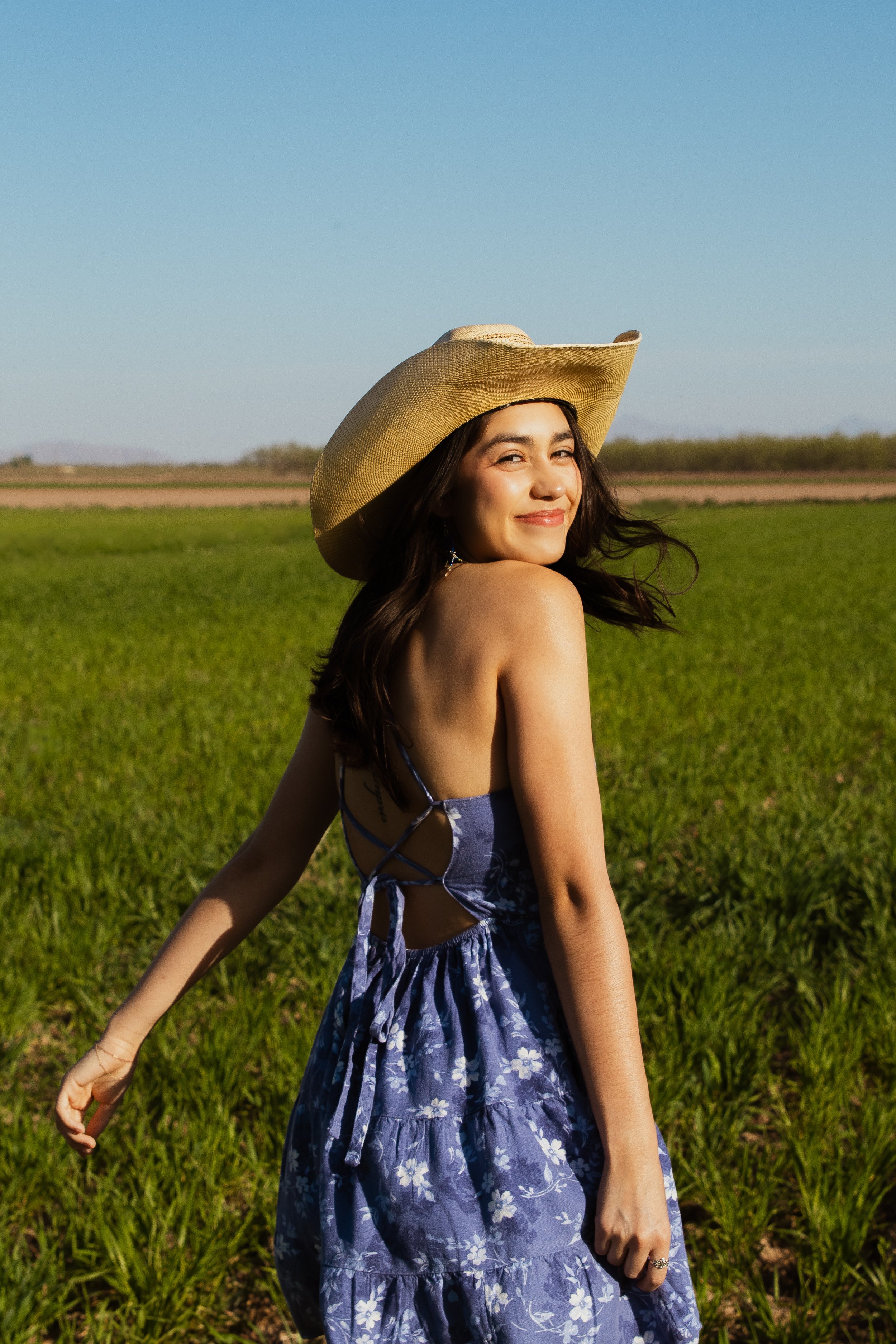 Young woman smiling in a meadow, wearing a large sun hat and a backless, floral purple dress.