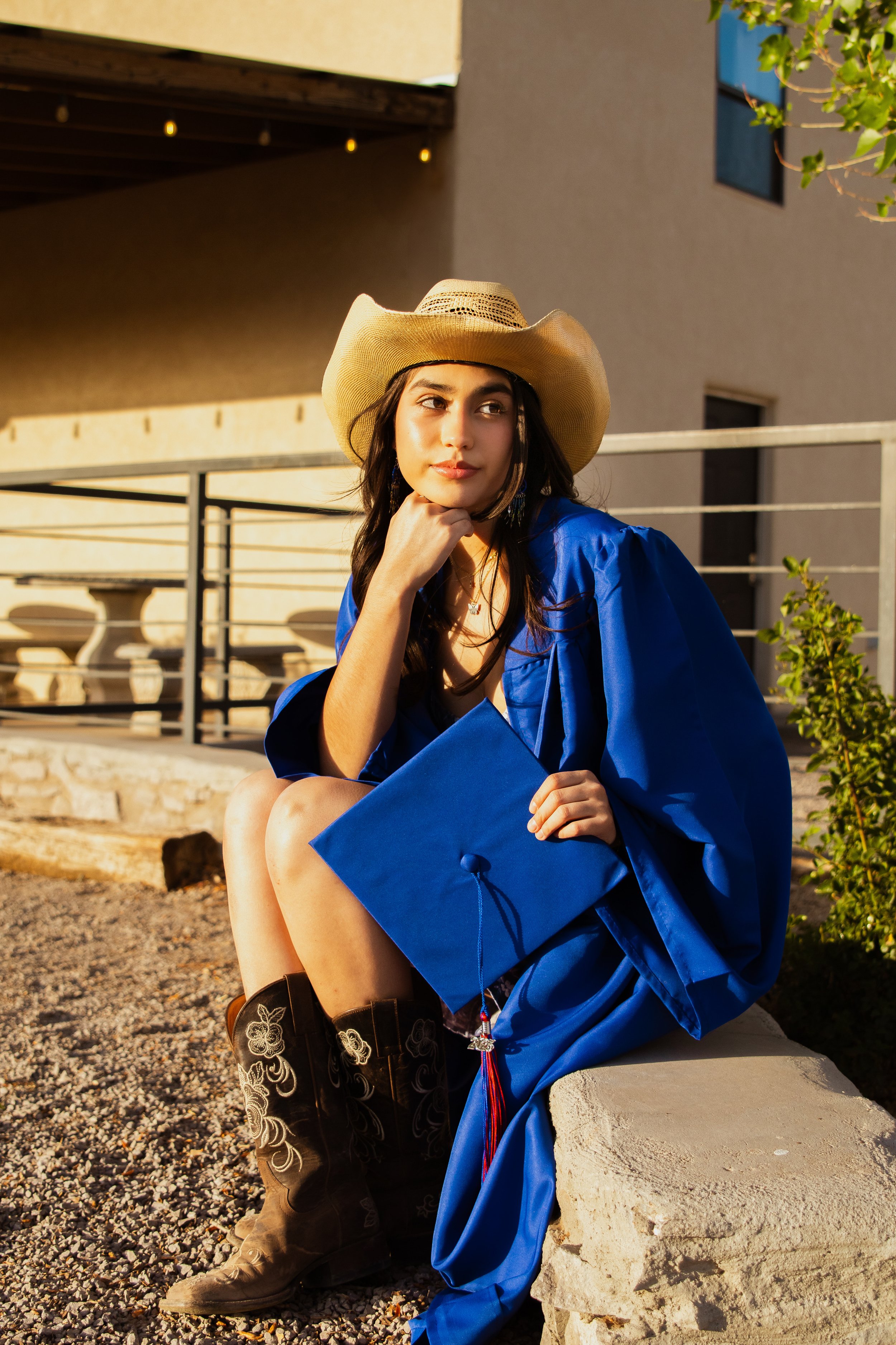 Young woman in a blue graduation gown, holding a cap, sitting on a stone bench outdoors, wearing a cowboy hat and boots, with buildings and greenery in background.
