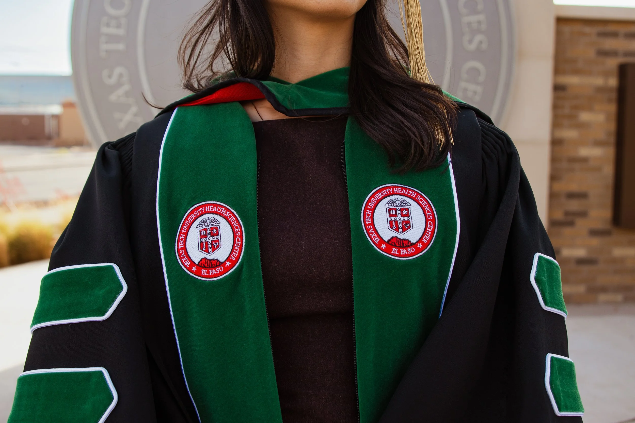 Person in black graduation gown with green and red accents, wearing a green stole embroidered with the Texas Tech University Health Sciences Center logo, standing outdoors.