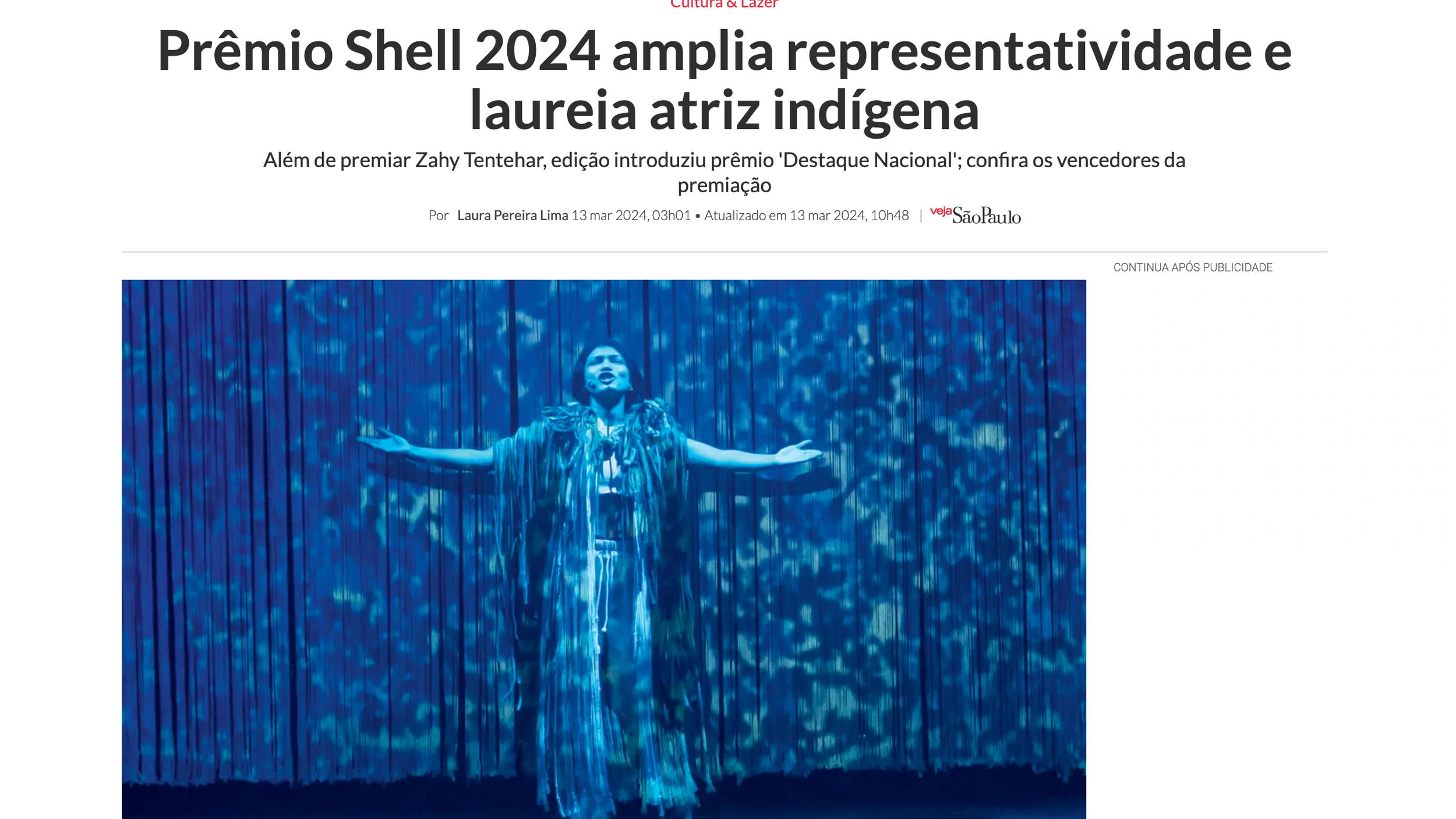 Imagem de uma mulher indígena em uma performance cênica, com braços abertos, vestida com roupas tradicionais, diante de um fundo iluminado com luzes azuis.