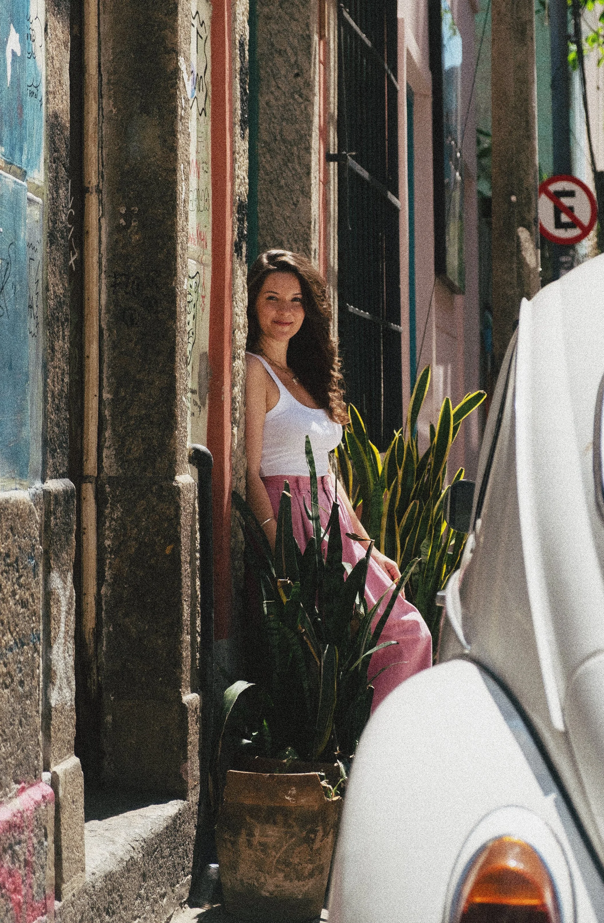 Yohana lopes. Mulher sorrindo sentada na rua, entre plantas e paredes com graffiti, parcialmente escondida por uma moto branca.
