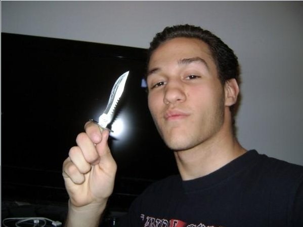 A young man with dark hair and a slight smirk holding a large silver knife in his right hand, standing in front of a television.