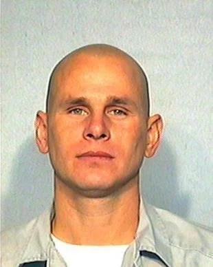 A bald man with a serious expression, wearing a light gray shirt, standing against a plain light-colored background.