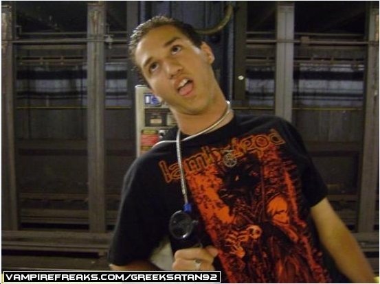 A young man with dark hair makes a funny face with his tongue out. He is wearing a black T-shirt with red graphic design, a necklace, and a stethoscope around his neck. The background shows metal racks and equipment.