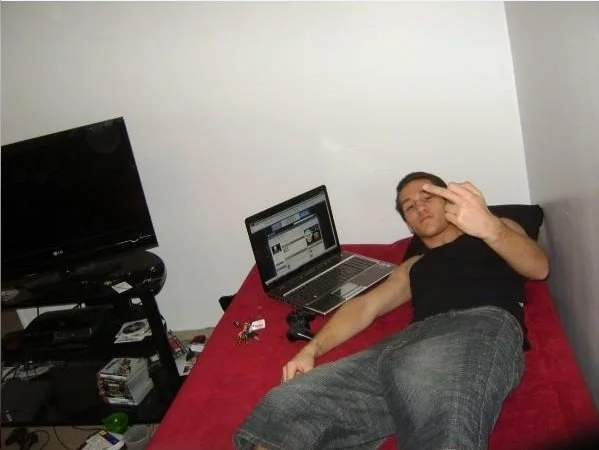 Young man lying on a bed with a red bedspread, using a laptop, next to a TV and entertainment stand with various electronic devices.