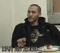 A young man with a shaved head wearing a black hoodie sitting at a table with a plate of food, looking to the side. The date March 25, 2009 is displayed.