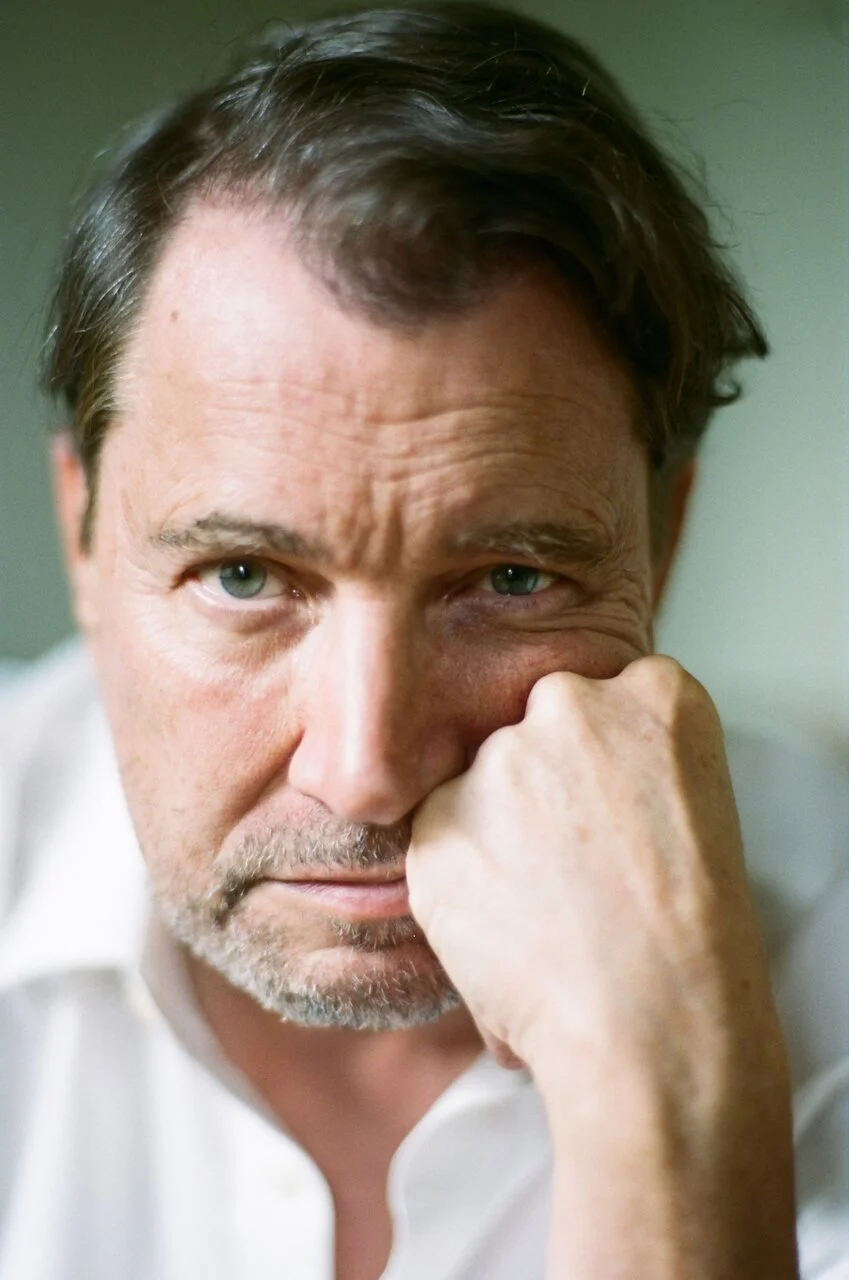 Close-up of a middle-aged man with green eyes and dark brown hair, resting his face on his hand, looking contemplative.