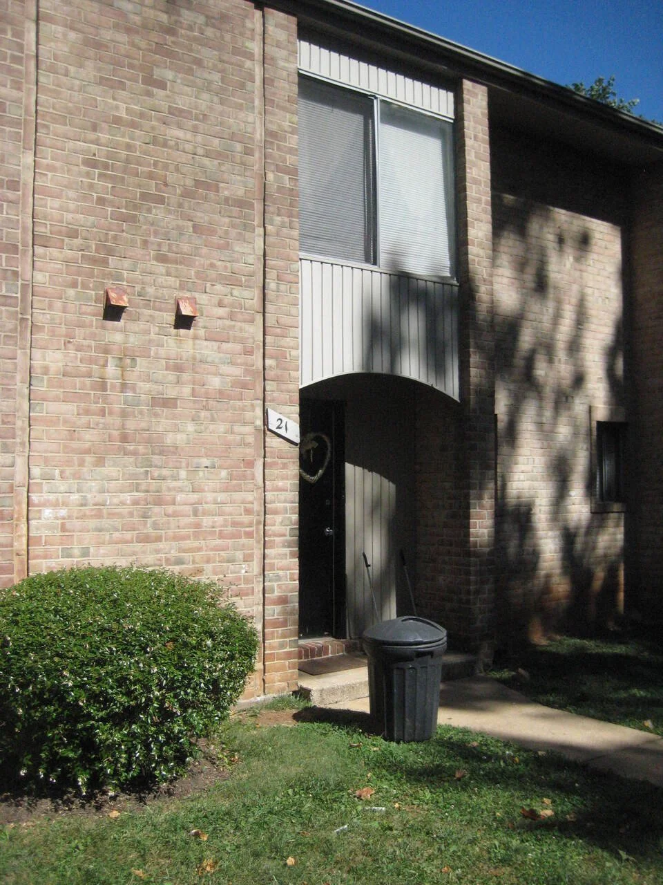 Brick apartment building entrance with black door, a window above, a bush, a trash bin, and tree shadows.