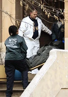 Two police officers and a man in white clothing near a staircase outside a building, with a large black bag and yellow caution tape.