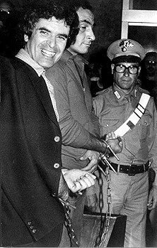 A black and white photo of three men, one speaking to a woman in military uniform, and another man in background, all in a casual indoor setting.
