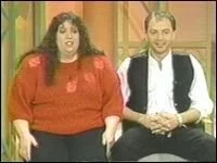 A woman with long dark hair wearing a red top and a man with short dark hair wearing a white shirt and black vest sitting together in a room with wood-paneled walls.