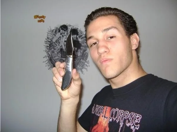 A young man with slicked-back hair holding a straight razor in front of his face, standing in front of a wall with a lion illustration and a small sticker that says 'Morganty'. he is wearing a black band t-shirt.