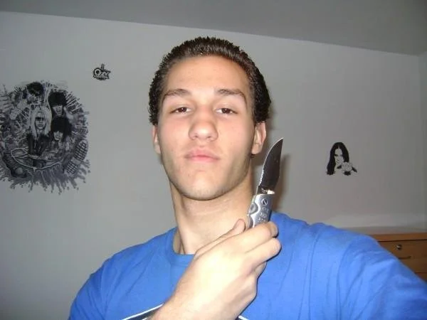 Young man with curly hair taking a selfie in a room, holding a knife near his face, with black and white posters on the wall behind him.