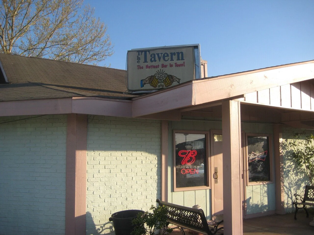 Exterior view of a bar called 'The Tavern' with a sign that says 'The Hottest Bar in Town!' above the entrance. The building has pink trim, a brick wall painted light green, and two windows with neon signs that say 'Budweiser' and 'OPEN.' There are benches and potted plants outside under a clear blue sky.