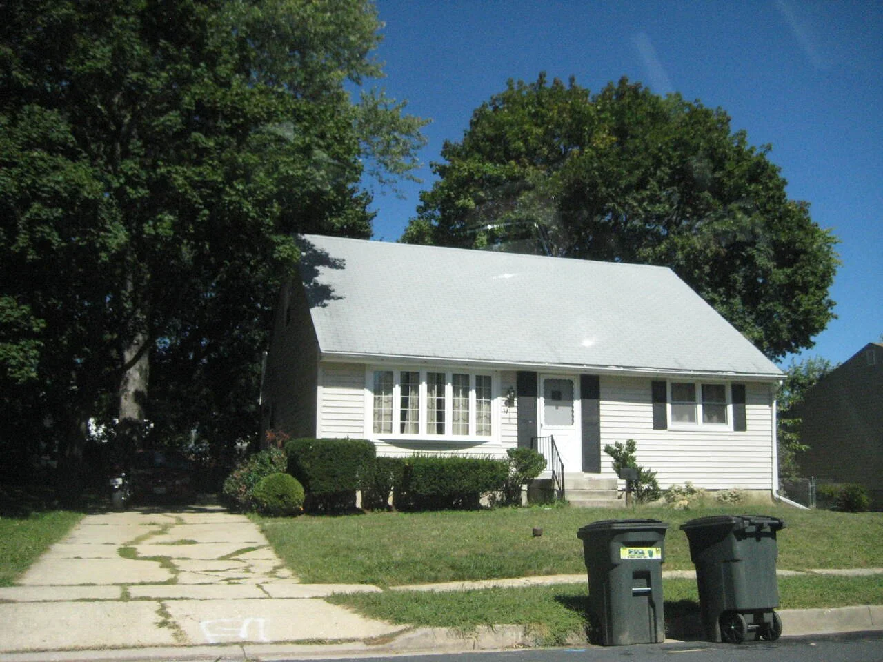 A small white house with a steep roof, bushes in front, two black trash bins on the street, and large trees in the background under a clear blue sky.