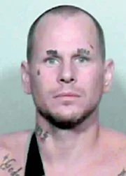 A man with a shaved head and facial tattoos staring at the camera against a plain, light-colored background.