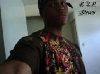 A young man taking a selfie indoors, wearing a black and colorful patterned shirt, with a light blue wall and a sign in the background.