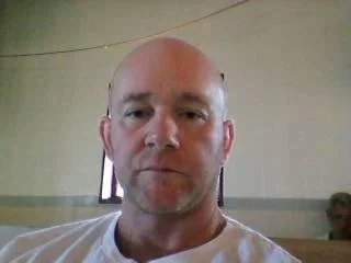 A close-up of a bald man with light skin, wearing a white shirt, in an indoor setting with a beige wall and a small television in the background.