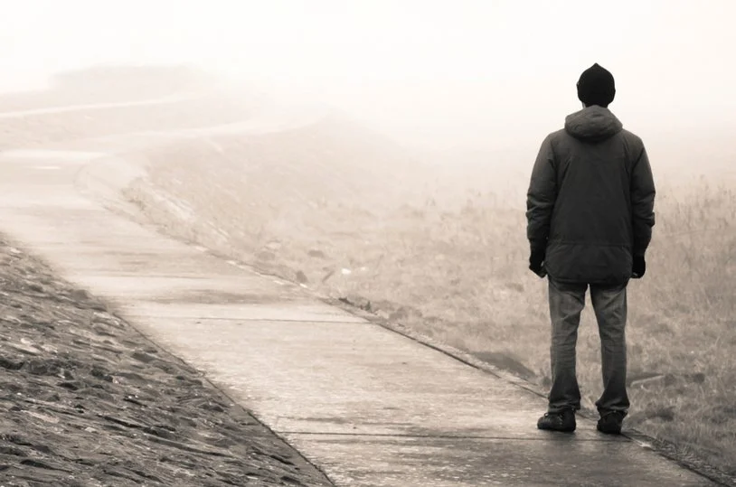 Person standing on sidewalk, facing away, in a foggy or misty outdoor setting
