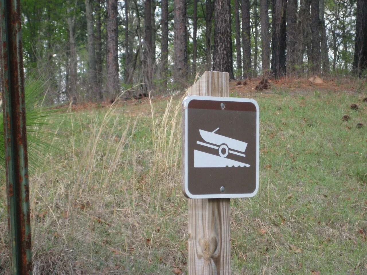 A sign with a graphic of a wheelbarrow, indicating a wheelbarrow crossing or caution area, mounted on a wooden post in a grassy outdoor area with trees in the background.
