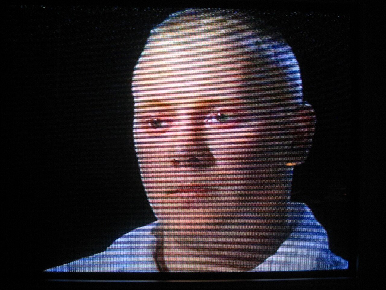 Close-up of a television screen showing a woman with short blonde hair and light skin, wearing a white shirt, against a black background.